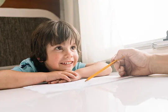 A young child smiling while engaging in an activity with an adult's hand, possibly drawing or writing with a pencil.