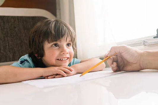 A young child smiling while engaging in an activity with an adult's hand, possibly drawing or writing with a pencil.