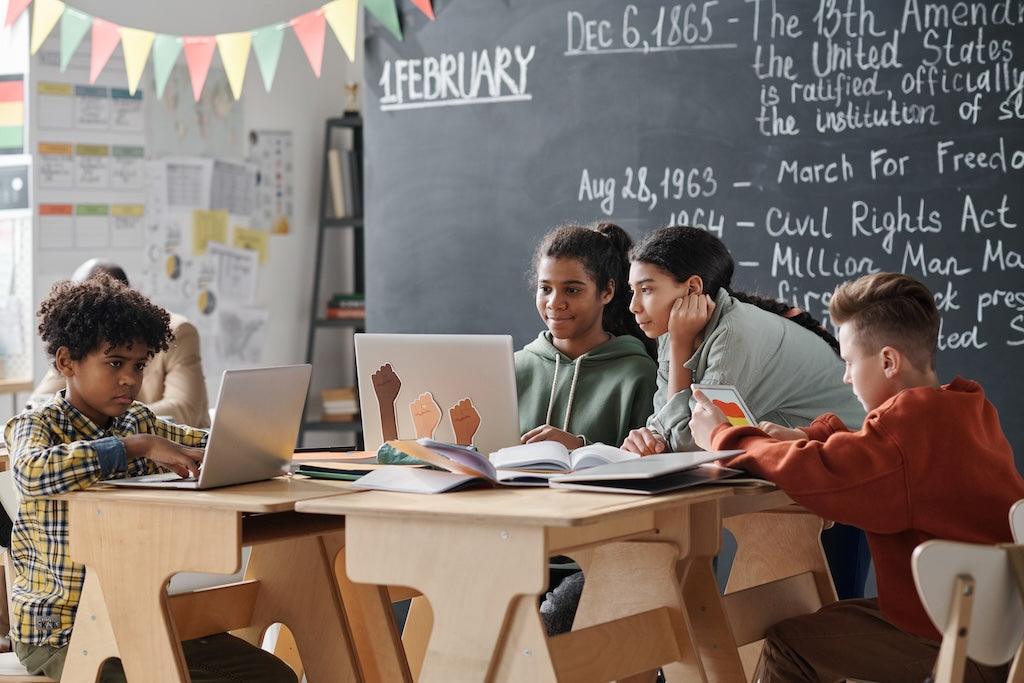 A group of students sitting around a table in a classroom setting, with laptops and notebooks, engaged in learning activities.